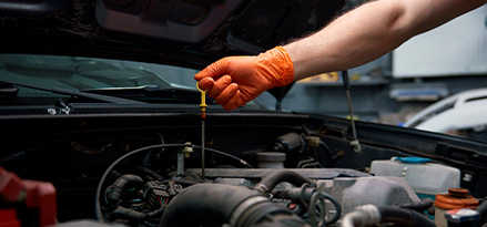 Mano con un guante naranja que sostiene una bayoneta de aceite para motor con restos de lubricante.