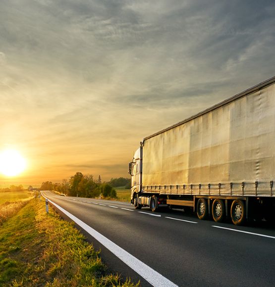 La imagen muestra un tractocamión con un remolque cubierto circulando por una carretera rural al atardecer, con campos abiertos a ambos lados y el sol bajo en el horizonte.