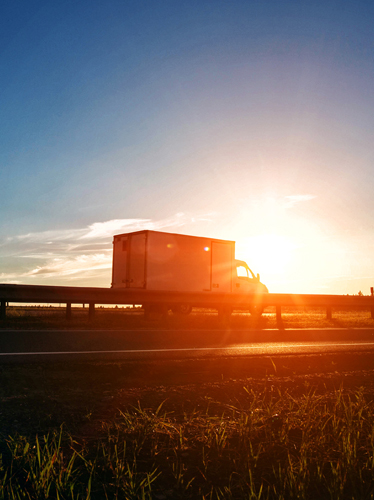 La imagen muestra un camión de caja circulando por una carretera al atardecer, con el sol bajo en el horizonte y el vehículo en silueta sobre un cielo iluminado en tonos anaranjados.